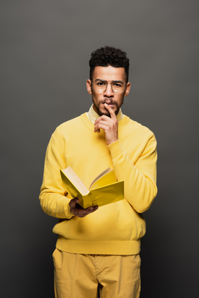 pensive african american man in glasses and yellow outfit holding book on dark grey - Photo, Image