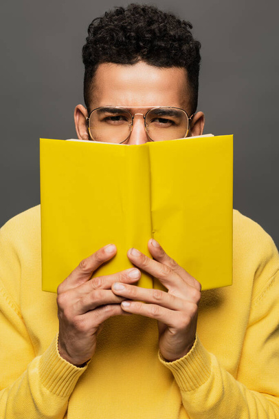 Afričan Američan muž v brýlích a žluté oblečení držení kniha izolované na šedé - Fotografie, Obrázek