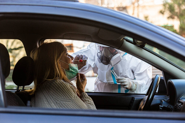 Pracownik medyczny wykonujący test drive-thru COVID-19, pobierający wymaz z nosa od pacjentki przez okno samochodu, diagnostyka PCR dla koronawirusa, lekarz w apteczce PPE. - Zdjęcie, obraz