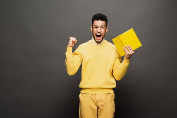 excité homme afro-américain en lunettes et tenue jaune tenant livre sur gris foncé - Photo, image