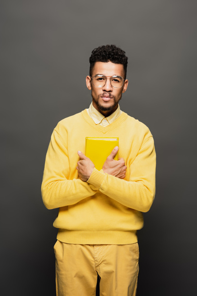 rizado africano americano hombre en gafas y amarillo traje celebración libro en gris oscuro - Foto, Imagen