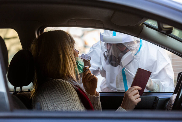 Medical worker performing drive-thru COVID-19 test, taking nasal swab sample from female patient through car window, PCR diagnostic, doctor in PPE holding test kit. She is holding passports. - Photo, Image