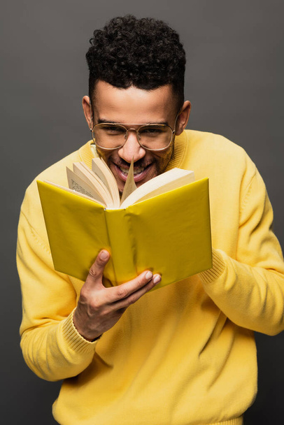 smiling african american man in glasses and yellow outfit reading book isolated on grey - Foto, imagen