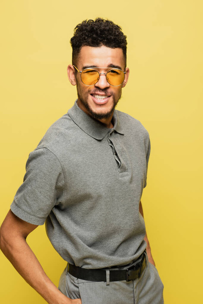 boldog afro-amerikai férfi napszemüvegben és szürke tenisz ing áll a kezét a zsebében elszigetelt sárga  - Fotó, kép