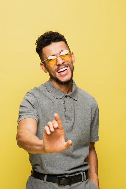 happy african american man in sunglasses and grey tennis shirt gesturing isolated on yellow  - Фото, изображение