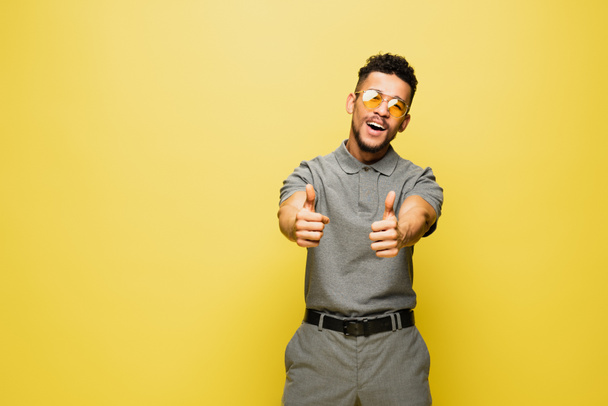 vreugdevolle Afro-Amerikaanse man met een zonnebril en een grijs tennisshirt met duimen op geel  - Foto, afbeelding