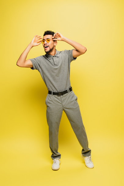 full length of cheerful african american man in grey tennis shirt adjusting sunglasses on yellow - Photo, Image