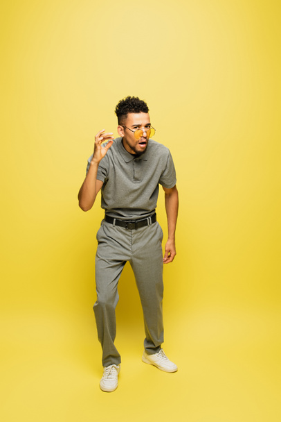full length of shocked african american man in sunglasses and grey tennis shirt standing on yellow - Foto, immagini