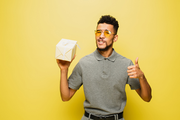 african american man in sunglasses and grey tennis shirt holding wrapped present and showing thumb up on yellow - Photo, Image
