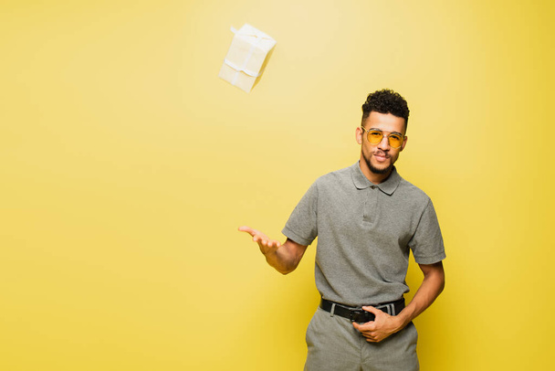 african american man in sunglasses and grey tennis shirt throwing wrapped present on yellow - Photo, Image