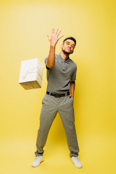 full length of stylish african american man in sunglasses and grey tennis shirt throwing wrapped present on yellow - 写真・画像