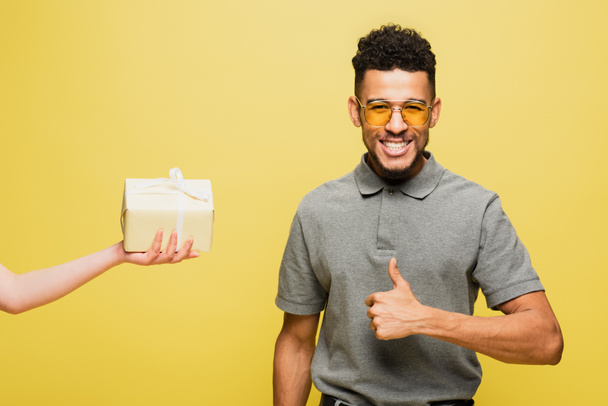 african american man in sunglasses showing thumb up near woman holding wrapped present on yellow - Foto, immagini