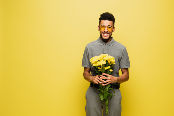 šťastný africký Američan v slunečních brýlích a šedé tenisové košili drží kytice květin izolovaných na žluté  - Fotografie, Obrázek