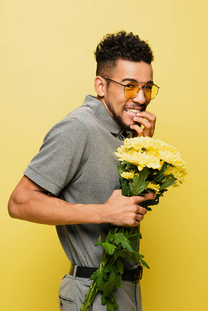 worried african american man in sunglasses and grey tennis shirt holding bouquet of flowers isolated on yellow  - Fotografie, Obrázek
