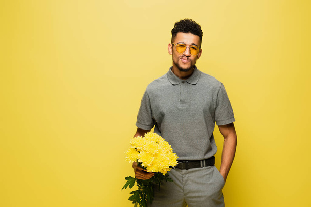 stylish african american man in sunglasses holding bouquet of flowers and standing with hand in pocket isolated on yellow  - Photo, Image
