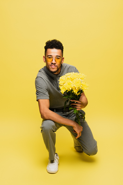 hombre afroamericano en gafas de sol sosteniendo ramo de flores y de pie sobre la rodilla en amarillo  - Foto, imagen