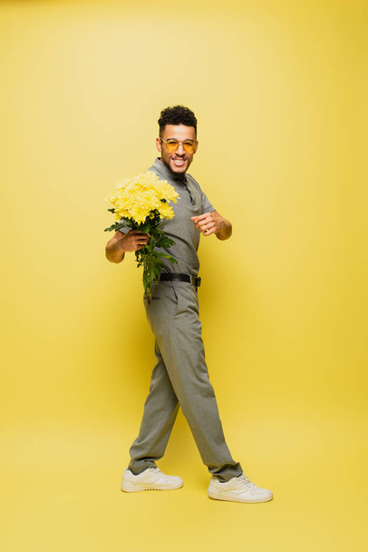 full length of happy african american man in sunglasses holding bouquet of flowers and standing on yellow  - Foto, immagini