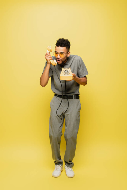 Güneş gözlüklü ve gri tenis tişörtlü Afro-Amerikan adam sarı telefonda konuşuyor.  - Fotoğraf, Görsel