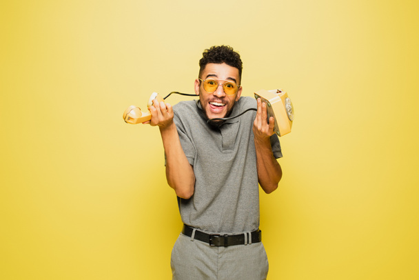 összezavarodott afro-amerikai férfi napszemüvegben és szürke tenisz ing kezében retro telefon sárga  - Fotó, kép