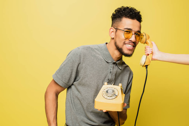 жінка тримає телефон біля веселого афроамериканського чоловіка в сонцезахисних окулярах і сірій тенісній сорочці на жовтому
  - Фото, зображення