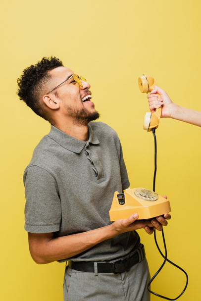 mujer sosteniendo el teléfono cerca del hombre afroamericano positivo en gafas de sol y camisa de tenis gris aislado en amarillo  - Foto, Imagen