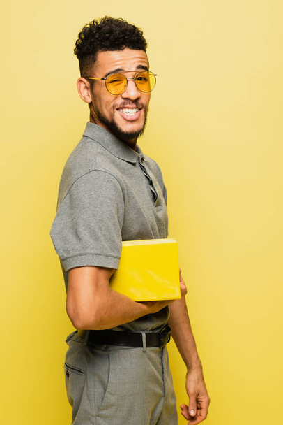 szczęśliwy Afroamerykanin człowiek w okularach przeciwsłonecznych i szarej koszuli tenisowej gospodarstwa książki izolowane na żółty  - Zdjęcie, obraz