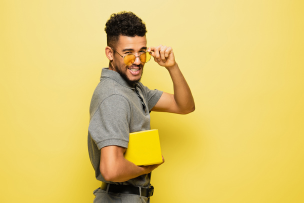 joven afroamericano hombre ajustando gafas de sol y sosteniendo libro en amarillo  - Foto, Imagen