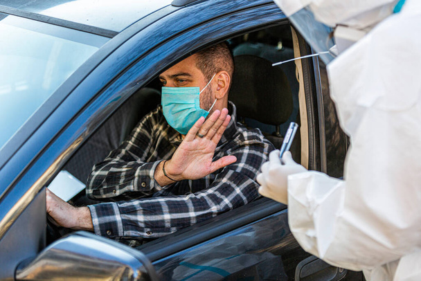 Man refuses medical worker trying to perform drive-thru COVID-19 test, taking nasal swab sample from patient through car window, PCR diagnostic, doctor in PPE holding test kit. She is holding passports. - Fotografie, Obrázek