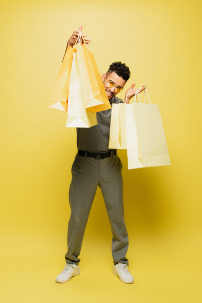 full length of positive african american man in sunglasses and grey tennis shirt holding shopping bags on yellow  - Φωτογραφία, εικόνα