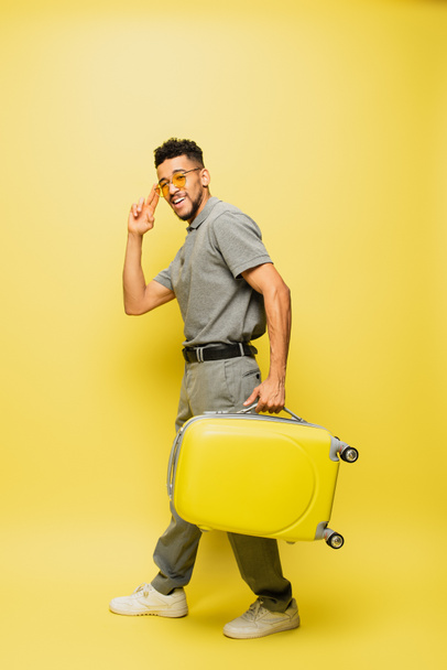 幸せなアフリカ系アメリカ人男性のサングラスとグレーのテニスシャツ黄色の上に荷物を運ぶ - 写真・画像