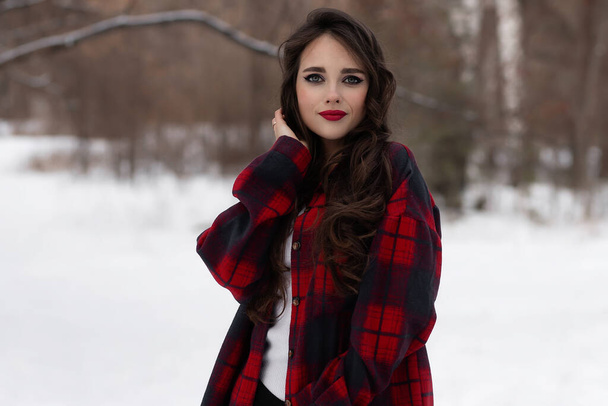 Urocza kobieta z czerwonymi ustami i w szachownicy dotykająca włosów stojąc w śnieżnym lesie zimowym i patrząc w kamerę - Zdjęcie, obraz