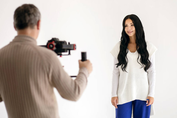 A beautiful brunette woman is vlogging with a cameraman in a white photo studio. Young woman blogger, journalist filming with a professional camera and videographer - Photo, Image