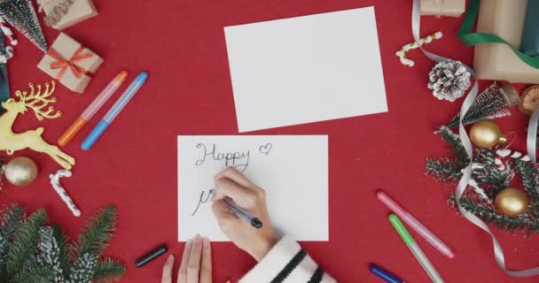 Top view of girl hands writing greeting card winter christmas decorations on red background. Minimal creative flat lay of christmas traditional composition and new year holiday season. - Footage, Video