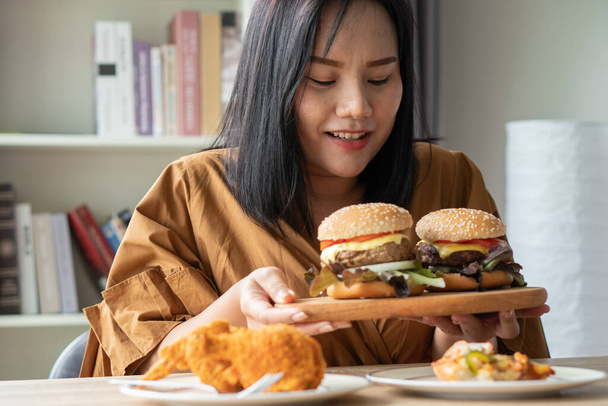 空腹太りすぎの女性は、木製のプレート上にハンバーガーを保持,テーブルの上にフライドチキンとピザ,自宅から仕事中,体重の問題を得る.大食障害の概念(BED). - 写真・画像