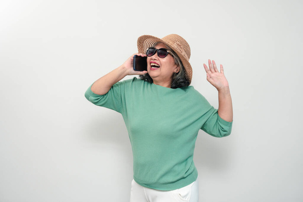 Portrait of happy asian senior woman wearing sunglasses, a hat, and holding smartphone standing isolated on white background. Concept of tourist enjoy and fun valentine day after retire - Photo, Image