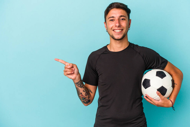 Nuori mies pelaa jalkapalloa pitäen palloa sinisellä taustalla hymyillen ja osoittaen sivuun, näyttäen jotain tyhjää tilaa. - Valokuva, kuva