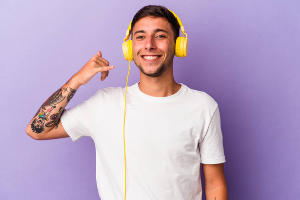 紫色の背景に孤立した音楽を聴く若い白人男性指で携帯電話のコールジェスチャーを示す. - 写真・画像
