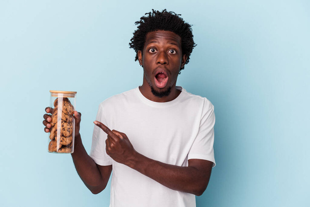 Young african american man holding chocolate chips cookies isolated on blue background pointing to the side - 写真・画像