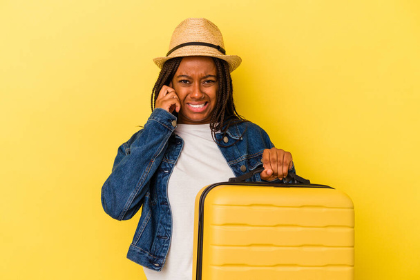 Молода африканка - мандрівниця, що тримає валізу ізольованою на жовтому фоні, закриваючи вуха руками.. - Фото, зображення