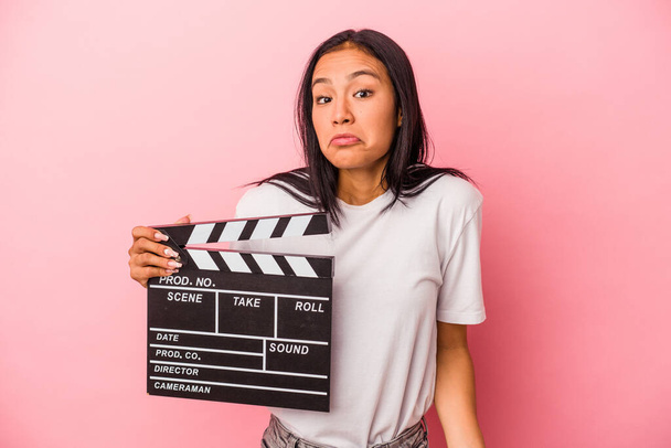 Young latin woman holding clapperboard isolated on pink background  shrugs shoulders and open eyes confused. - Photo, Image