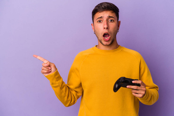 Giovane uomo caucasico in possesso di controller di gioco isolato su sfondo viola che punta al lato - Foto, immagini