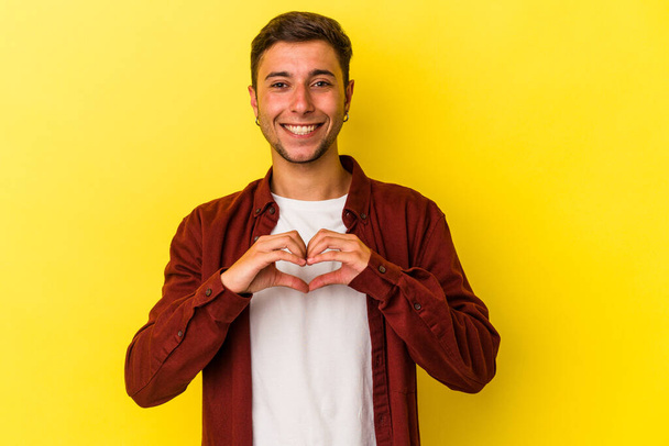 Jovem caucasiano com tatuagens isoladas em fundo amarelo sorrindo e mostrando uma forma de coração com as mãos. - Foto, Imagem