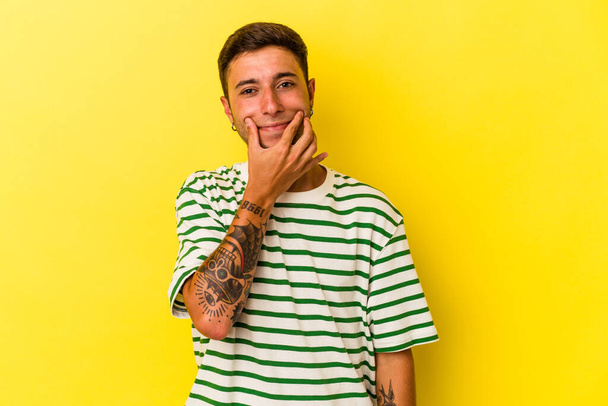 Joven hombre caucásico con tatuajes aislados sobre fondo amarillo dudando entre dos opciones. - Foto, imagen