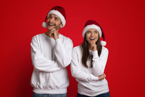 Pensive mladý pár na sobě Santa klobouky přemýšlet o něčem přes červené pozadí - Fotografie, Obrázek