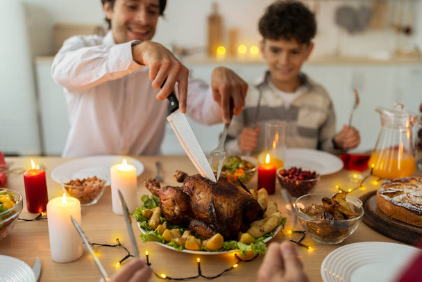 Gelukkig vader snijden geroosterde kerst kalkoen voor zijn zoon en familie op feestelijke geserveerd tafel binnen, selectieve focus - Foto, afbeelding