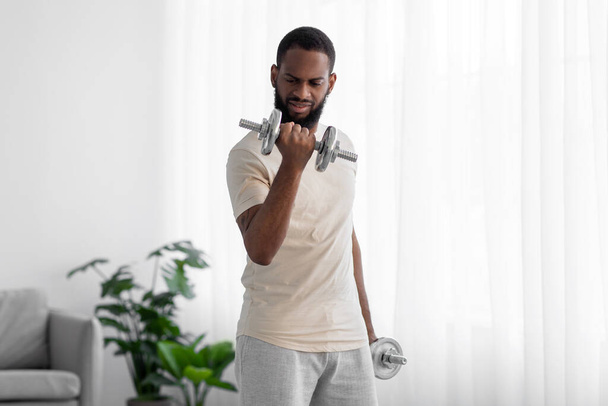 Strong young black male with beard in white sportswear raises dumbbell in room interior on window background - Fotoğraf, Görsel
