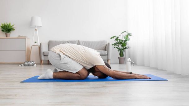 Calm young african american male with beard in white sportswear practicing yoga at floor at mat - Photo, Image