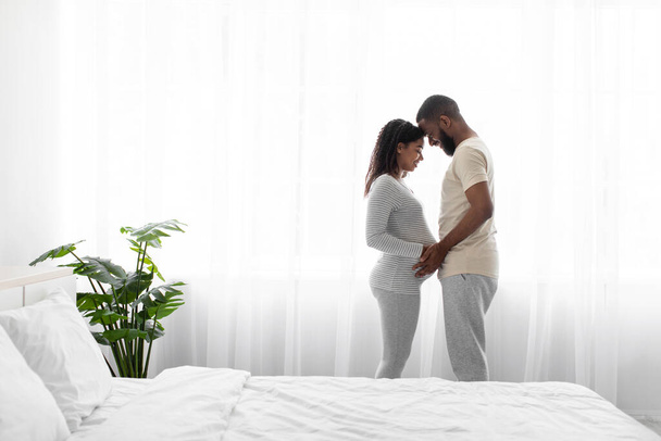 Vrolijke jonge zwarte man en zwangere vrouw genieten van tedere moment in slaapkamer interieur op venster achtergrond - Foto, afbeelding