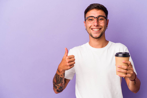 Junger kaukasischer Mann hält Kaffee zum Mitnehmen isoliert auf lila Hintergrund lächelnd und hebt den Daumen - Foto, Bild