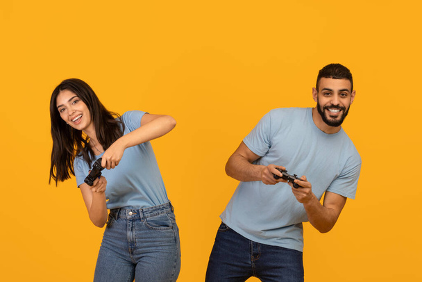 Η έννοια του τρόπου ζωής. Χαρούμενο ζευγάρι arab που παίζει online βιντεοπαιχνίδια, κρατώντας gamepad joystick, κίτρινο φόντο - Φωτογραφία, εικόνα